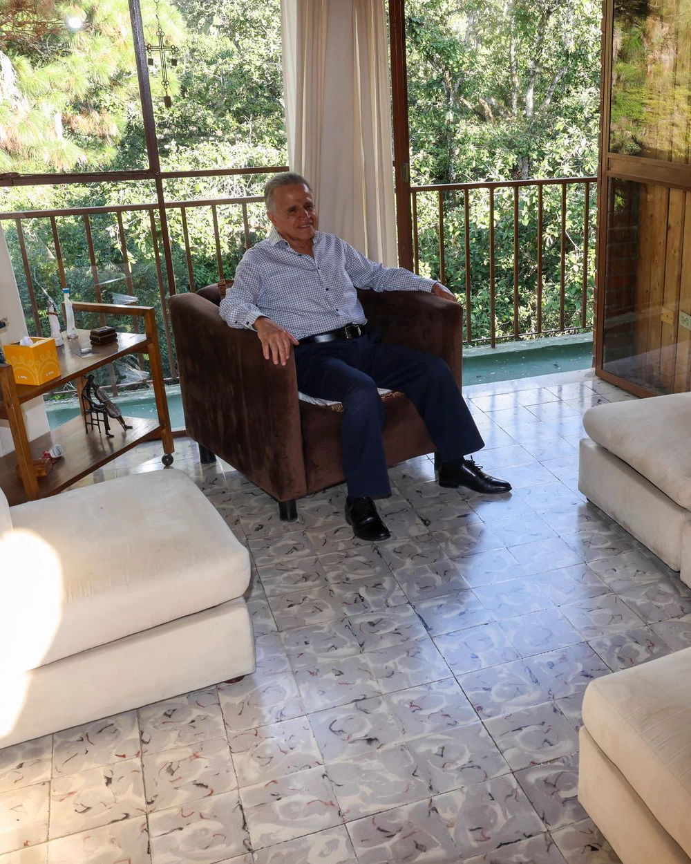 Lic. Fernando Estrada sentado en un sillón junto a un ventanal, con el follaje del balcón de fondo